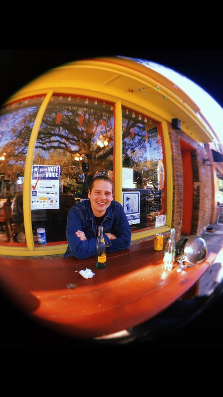 Writer Isaac George Lauritsen, wearing a jean jacket with hair pulled back, sits at a picnic table outside Panchita's in New Orleans, smiling.