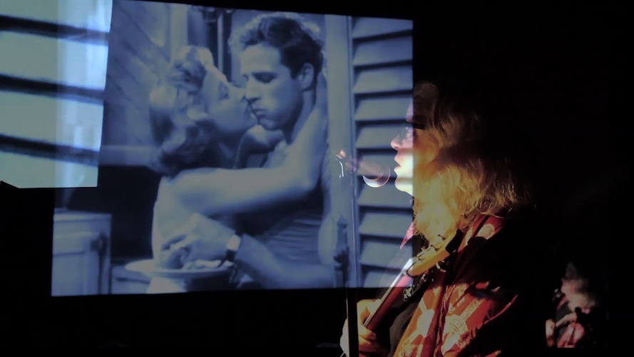 Neo-Benshi performer, Roxi Power, stands to the right of the screen, speaking into a microphone. She narrates new words spoken by the characters, Stanley and Stella Kowalski, from A Streetcar Named Desire.  Stella is kissing Stanley on the cheek as he eats from a plate. Photograph by Marilyn Rivchin.