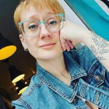 white femme with short blonde hair, large aquamarine glasses, and a prominent arm tattoo gazes wistfully at the camera