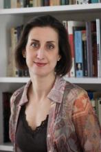 A woman with dark brown hair and pale skin poses in front of a bookcase wearing a brown shirt with silk blouse on top.