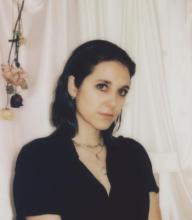 they/she with dark brown hair sitting in front of a silk background wearing a black shirt