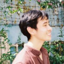 Damon in profile view, against a sky-blue wall with pink flowers and green leaves. He wears a dark red shirt and is smiling.