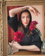 a dark haired woman gestures within a golden frame, two red carnations point towards and away from her