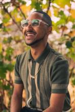 A photo of poet DeeSoul Carson. He is seen smiling in a green shirt with foliage around him