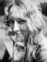Black and white close-up of a woman with wavy hair partially covering her face. She smiles, resting her hand on her chin.