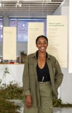 Picture of author wearing green jacket and matching cargo pants, a black shirt, and crystal necklace, smiling and facing the camera directly, and with poems hanging from ceiling in background 