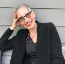 Author photo of Sarah Rosenthal from the waist up, with short black-and-white hair, glasses,  pink lipstick, a pink bead necklace, black shirt, and black sweater, leaning head on right hand and smiling into the camera.