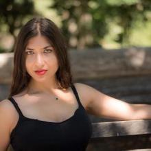an image of a non-binary person with long dark hair, olive skin, and green eyes in a black tank top sitting on a bench