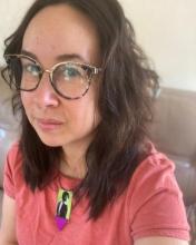 photo of a woman with brown medium-length wavy hair, wearing glasses, a pink tee shirt, and a necklace with a picture of Iranian poet Farrokh Farrokhzad