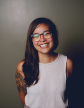 Danni is an Asian American poet with glasses and long dark hair. She smiles in front of a gray background.