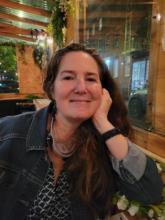 A picture of poet Sara Wallace sitting at an outdoor restaurant in New York. She is wearing a jean jacket, has long hair, and is smiling.