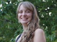 A white woman with long dark blonde hair smiles in front of a background of blurred green leaves.