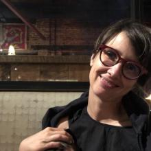 The image shows a person with short dark hair and wearing large, round, red eyeglasses. They are smiling.