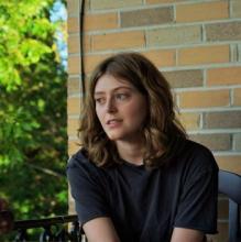 A young white woman with wavy brown hair sits in front of a light brick wall, looking out slightly left of camera. 