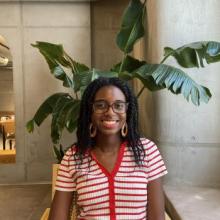 A photo of the poet Jasmine Flowers smiling and sitting in front of a monstera plant in a cafe with concrete walls.