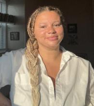 A person with tan skin, brown eyes, freckles, and blonde box braids faces the camera smiling. She is wearing a white button down shirt. 