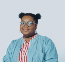 Portrait of a black woman from the chest up, wearing a blue denim shirt over a red and white striped dress. Woman is wearing glasses with short natural hair styled into two small space buns.