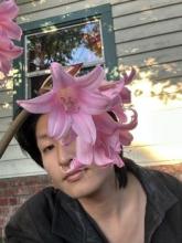 Figure in black jacket poses behind a branch of pink lillies.  