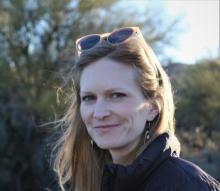 Kelsi is a white woman with blue eyes and long dark blonde hair held back by her sunglasses perched on her head. She looks sideways at the camera, smiling slightly.