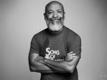 Black & white photo of a Black man smiling with his arms crossed, wearing a Song of Solomon tshirt and full white beard.