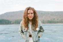 A person with curly red hair wearing a jean jacket standing in front of a lake