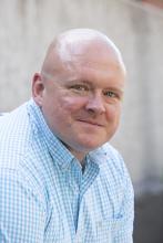 The photo shows Steve Halle, who is a white man with a shaved head wearing a blue and white plaid shirt.
