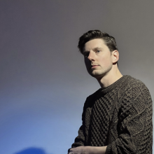 William Hazard wearing a sweater in front of a projector screen with blue and yellow lights shining on it