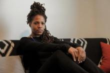 Black person with locs in a high bun and over shoulder, dressed in black pants and shirt on a couch, smiling slighty. 