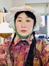 Stine is staring straight into the camera with a deadpan expression. She is wearing a plush white and green daisy flower hat that has a green chinstrap like a helmet. Her hair is in two braids. She is wearing yellow eyeshadow and a windbreaker with a purple camo pattern.. 