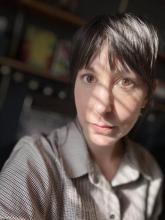 Middle-aged white feminine person with dark hair wearing a fawn blouse with sun through blinds on face