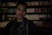 Taken via laptop, I'm wearing a black shirt and a black and gray keffiyeh, sitting in front of a bookshelf.