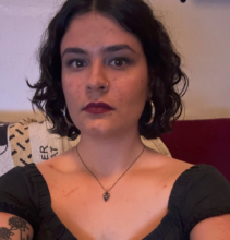 A South Asian woman with chin-length black hair in a black top and silver pendant necklace sits on a dark red sofa.