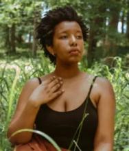Destiny Hemphill with a short cropped afro. They wear a black bodysuit and are seated in grass. Her head is turned to the right and is lightly tilted up.