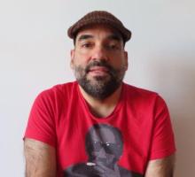  A headshot of a light-skinned, middle-aged, male-presenting, mixed-race Puerto Rican with facial hair wearing a brown flat cap and a red t-shirt with an image of the writer Steve Cannon