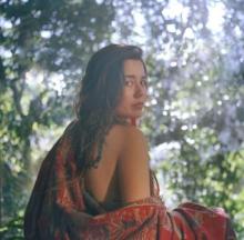 brown haired woman draped in red shawl looks over her right shoulder. behind her are trees. 