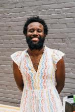 JJJJJerome stands and smiles in front of a white brick wall. He wears a white dress with yellow, light-blue, pink-orange, beige, and white flowers embroidered on it. He wears a gold chain and hold his hands behind his back. His skin is brown and his hair and beard are black. The sun shines on his face.