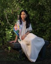 Headshot of Michelle Phương Hồ, who is sitting on a bench in a white dress with a colorful scarf.